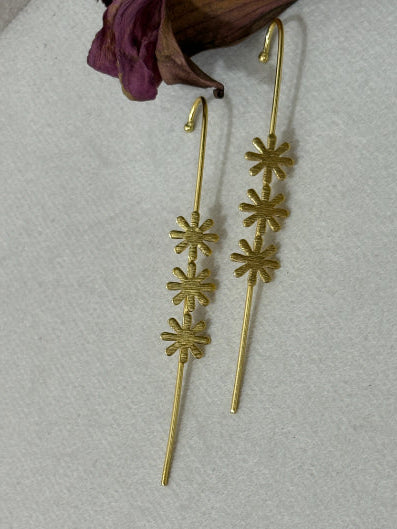 A pair of gold-toned barrette hairpins with three textured starburst motifs on each pin, displayed on a light gray textured surface with a dried purple flower prop in the corner.