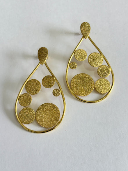 A pair of gold teardrop-shaped dangle earrings with textured circular cutouts in various sizes, displayed on a light off-white background.