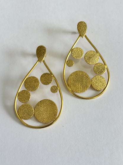 A pair of gold teardrop-shaped dangle earrings with textured circular cutouts in various sizes, displayed on a light off-white background.